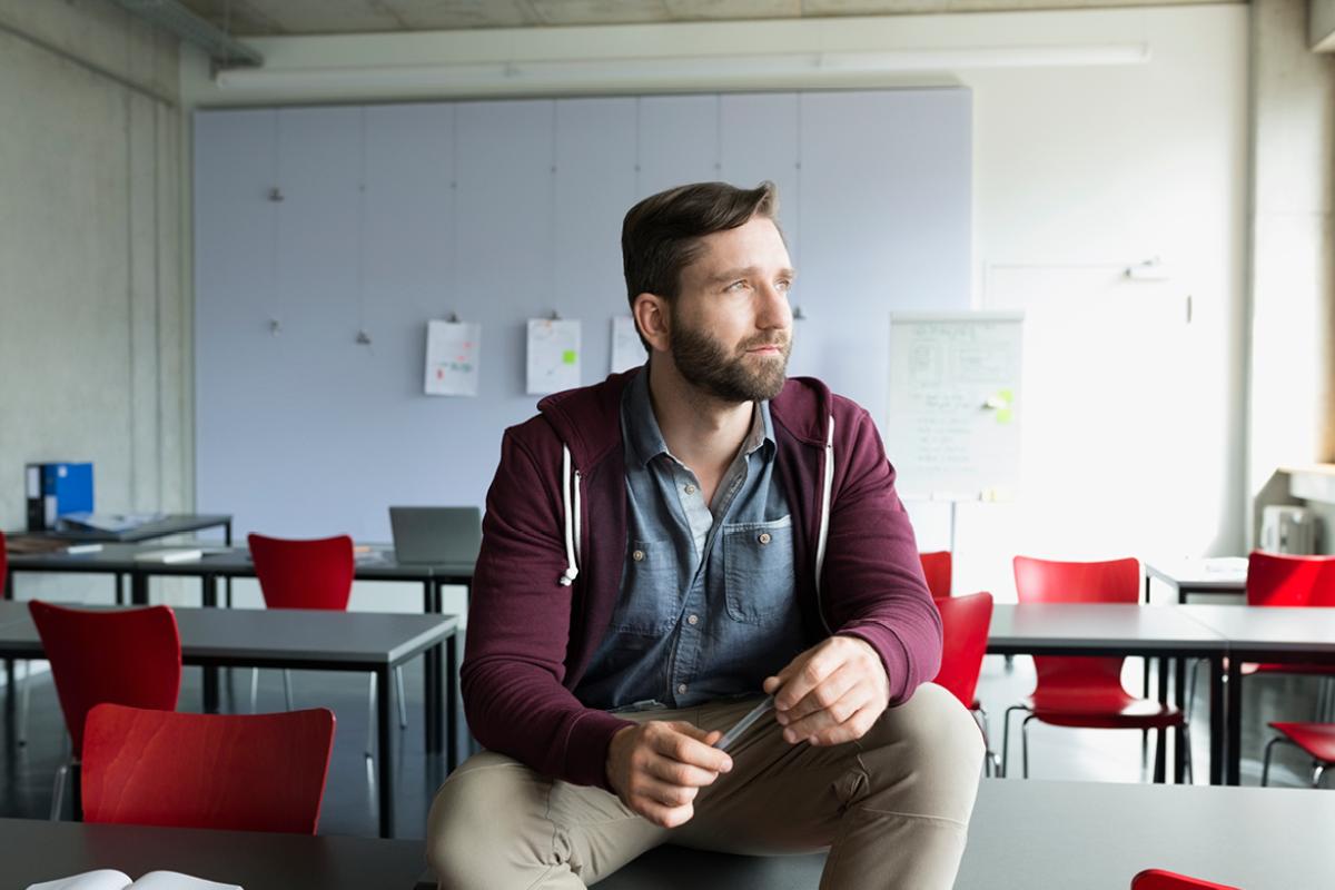 A man sitting on a table in an empty classroom holding a pen and looking out the window.